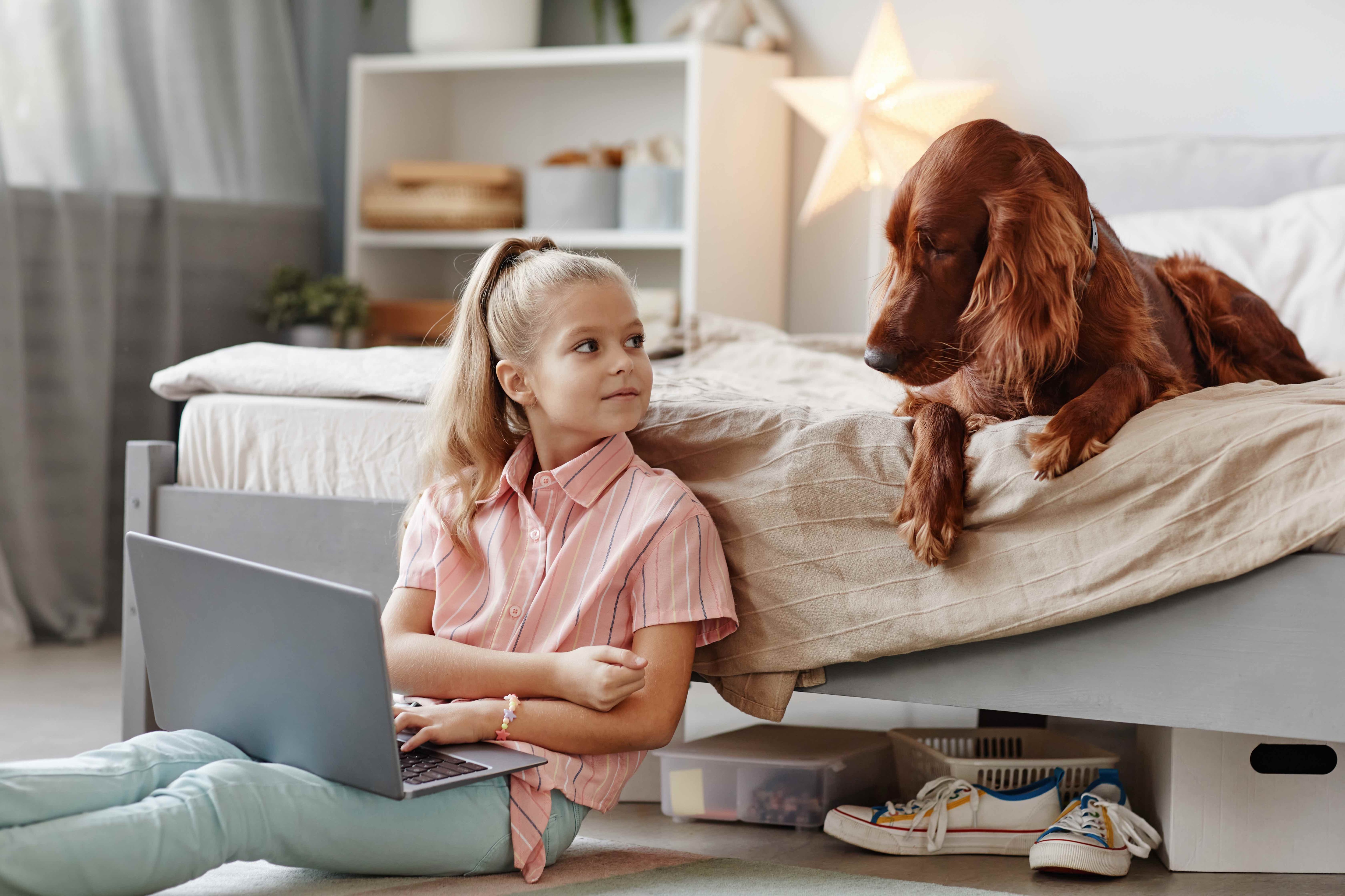 little girl sitting on her bedroom floor with an irish setter lying on her bed