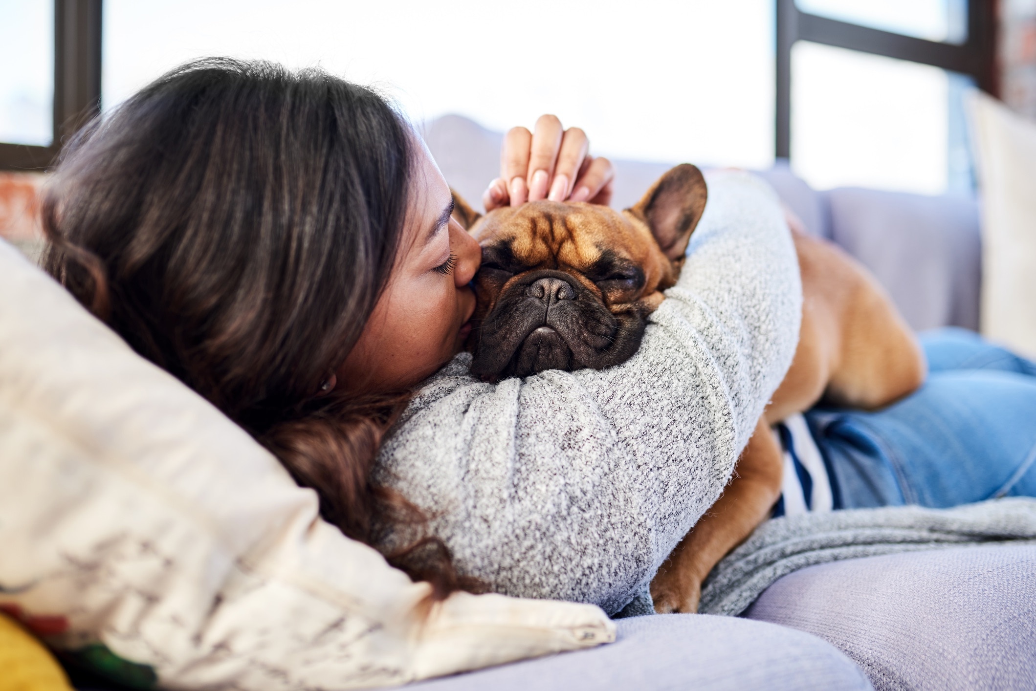 woman lying on the couch, hugging and kissing a brown french bulldog