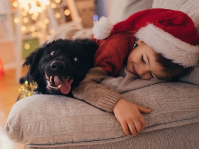 little kid in santa hat cuddles with black dog during the holidays