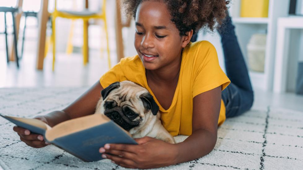 little girl reads book to pug while lying on floor
